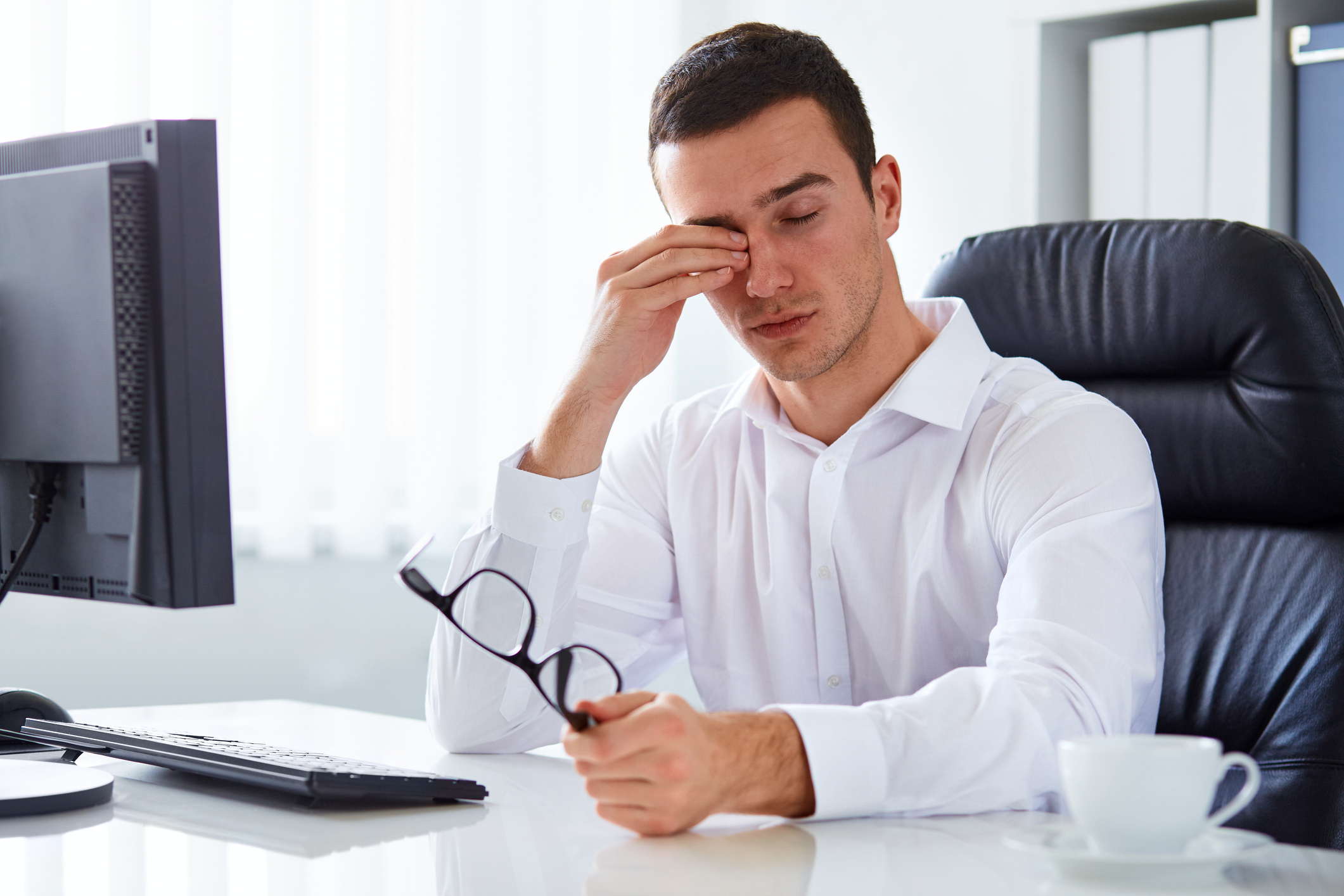 A man seated at a desk in front of a computer holds his glasses and rubs his eyes.
