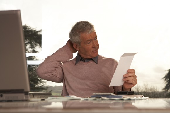 A senior man sitting at a table scratches his head as he looks at a bill.