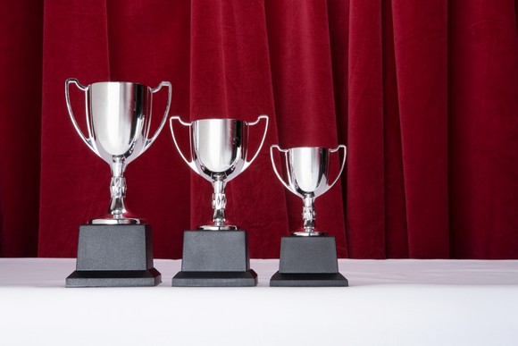 3 silver trophies of descending size, with a red curtain behind them.