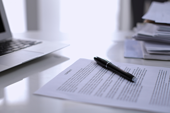 A laptop, pen, and paper contract sitting on a desk.