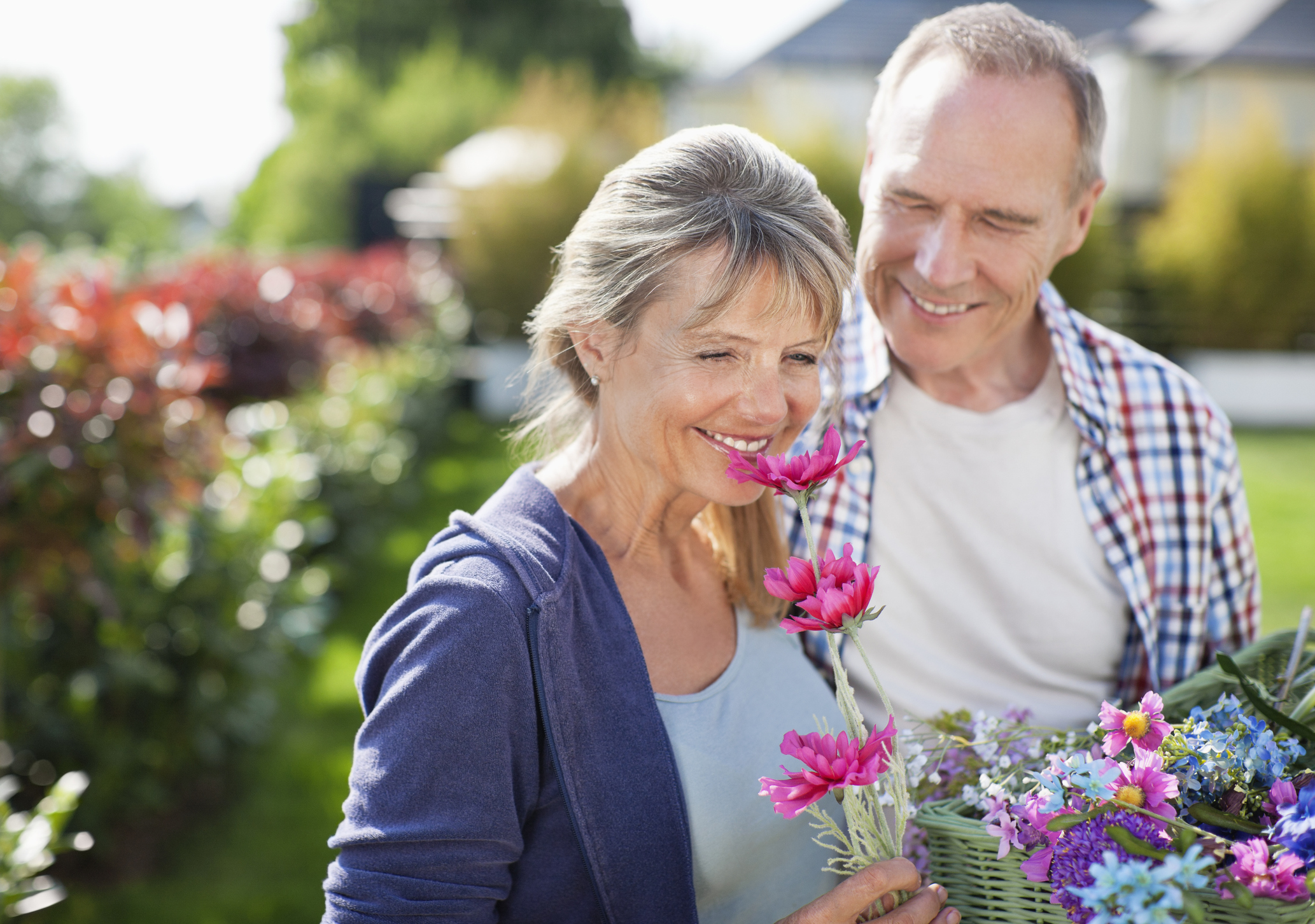 Senior woman smelling flowers and smiling while standing next to a smiling senior man.