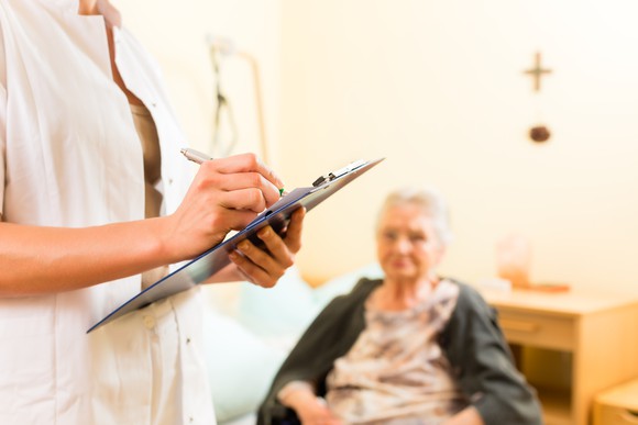 Elderly woman seated in the background, with a person in a lab coat holding a clipboard and pen in the foreground.
