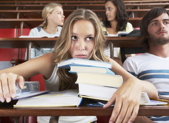 College student in lecture hall with chin on stack of textbooks, looking overwhelmed.