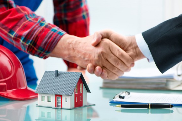Two hands in a handshake, one arm with a suit, other with a flannel shirt, with hardhat, toy house, and piles of papers on the desk below them.