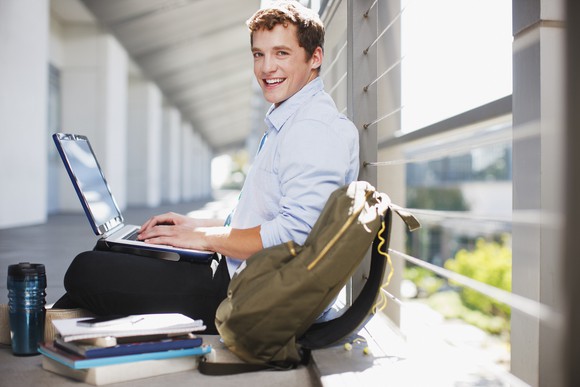Smiling young man typing on laptop with backpack and pile of books next to him.