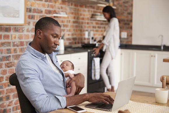 man using laptop computer while holding baby while woman cooks in background