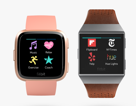 The Versa on the left and Ionic on the right. Versa is slightly smaller and has rounded corners, displayed in pink. The Ionic is in gray and has a brown leather wristband.