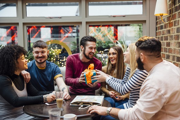 Young adults sitting around a table giving one of them a gift.