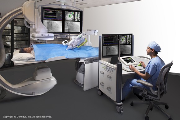 A seated physician in scrubs using the CorPath GRX System with a patient lying under it