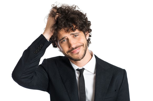 A confused, lightly bearded young man in a suit scratching the top of his curly hair.