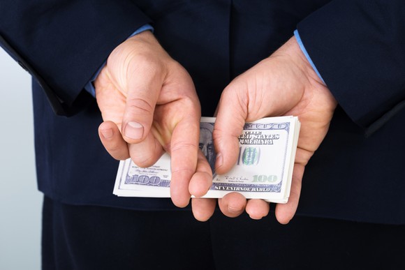 A businessman in a suit with his fingers crossed holding a stack of cash behind his back.