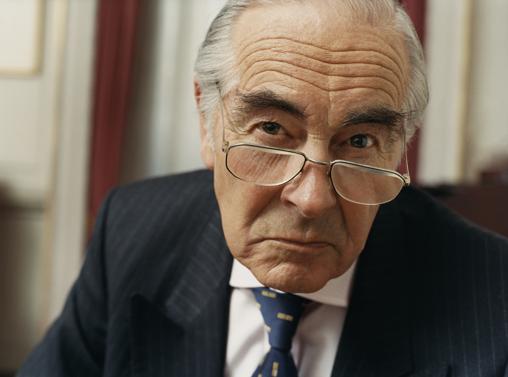 A wealthy older man in a suit scowling.