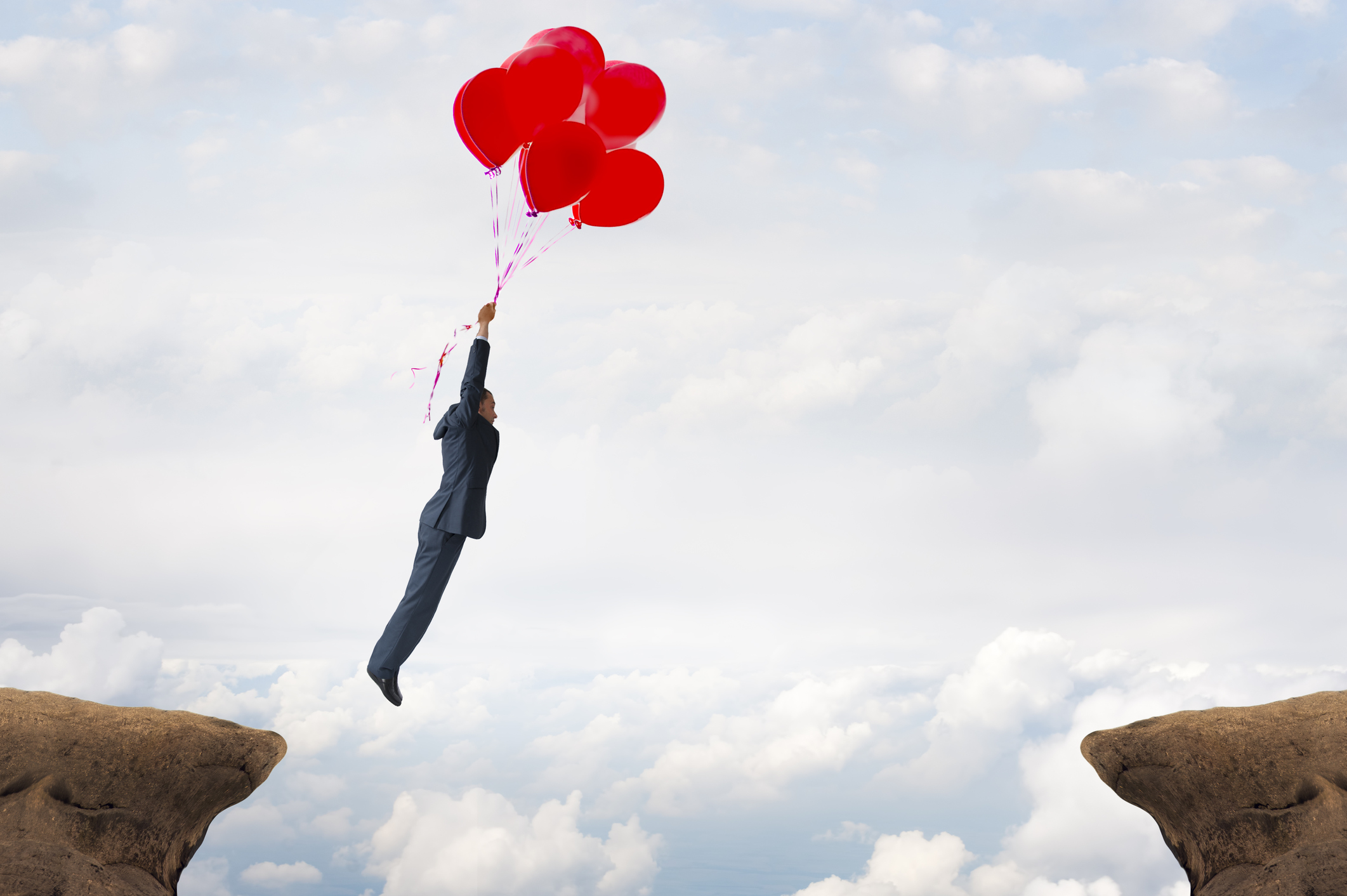 A man in a suit being carried across a wide gulf by ballons.