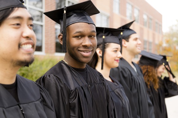 Row of smiling young adults in black graduation robes and hats standing outside.