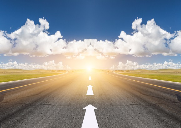 A road with arrows pointing forward towards a bright sun.