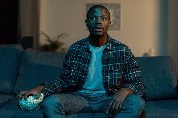 A man sitting on a couch, watching TV and eating popccorn.