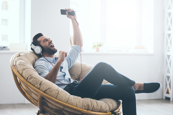 Man sitting in papasan chair listening to music on his smartphone via headphones