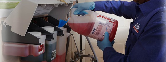 Worker wearing Cintas uniform pouring red liquid into a dispenser of multiple liquids.