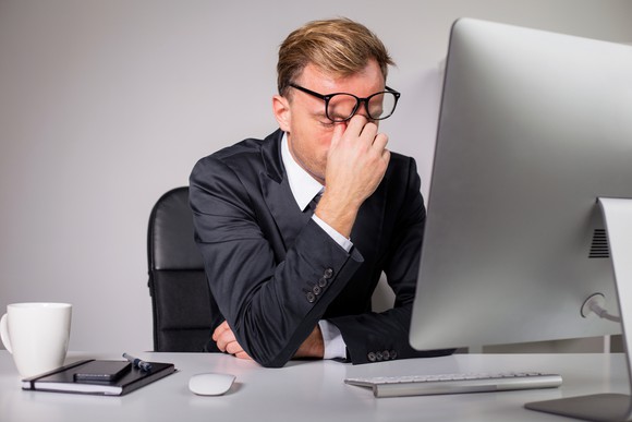 Man in suit with eyes closed while sitting at computer and right hand pinching forehead