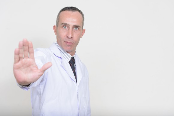 A man in a lab coat holds his hand up with the palm facing out