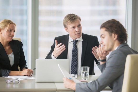 Man in suit gesturing toward another man in suit with an angry expression while a professional woman looks on.