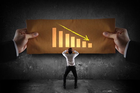A stressed-out businessman with both his hands on the back of his head while looking at a falling graph being held by another pair of hands.