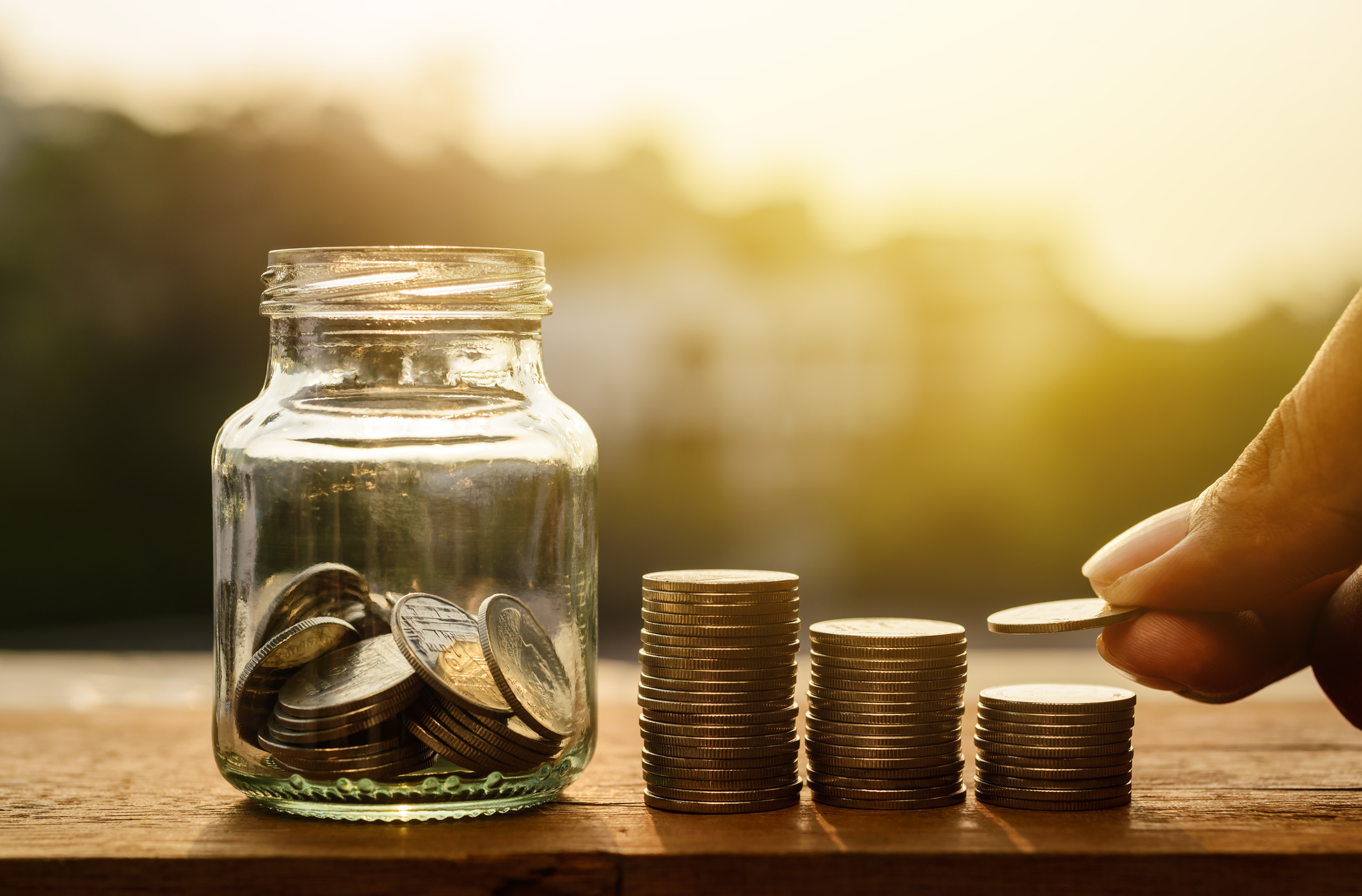 A hand putting money from rising coin stacks into a glass jar.