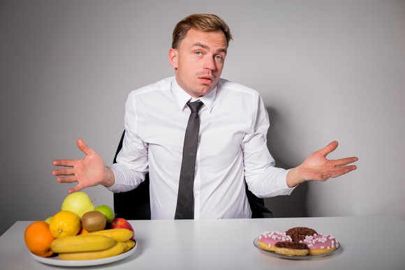 Man in white shirt shrugging over his dining choices -- a plate with four donuts or one with fresh fruits piled up