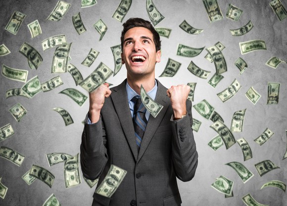 A business man pumping his fists in excitement as dollars rain on him against a grey background.