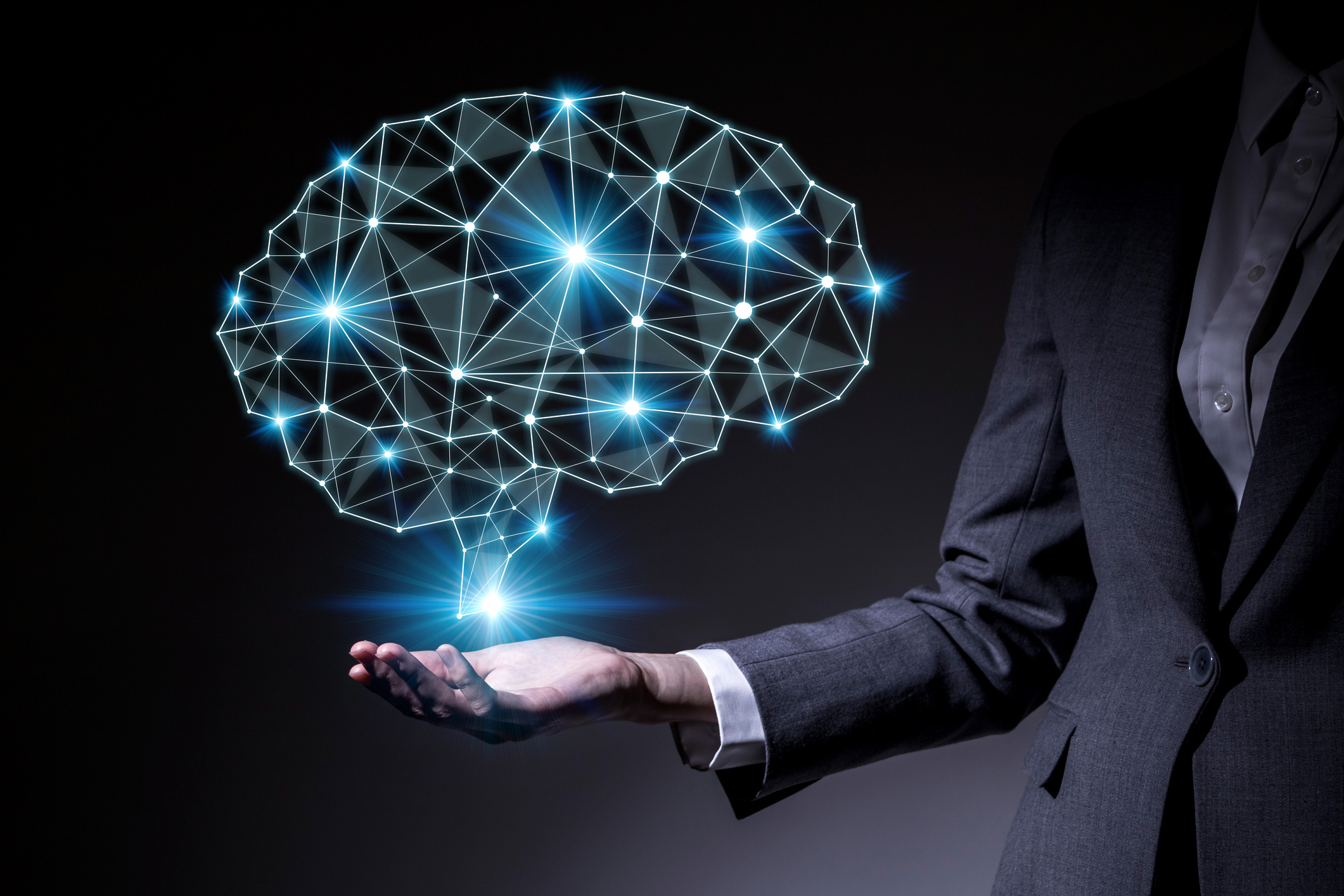 An image of a digital brain hovering above the right palm of a woman who is dressed in a business suit and holding her right hand out. Concept for AI.