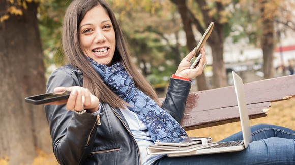 A woman sitting on a park bench and holding a laptop, a tablet, and three smartphones.