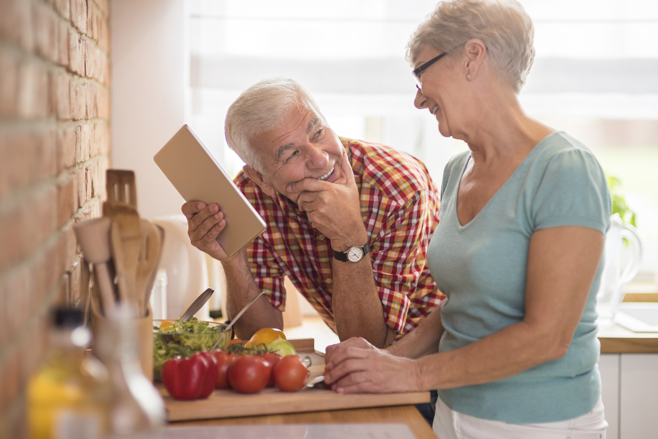 Senior man holding a notepad and smiling up at a senior woman while standing in a kitchen as she's cutting vegetables