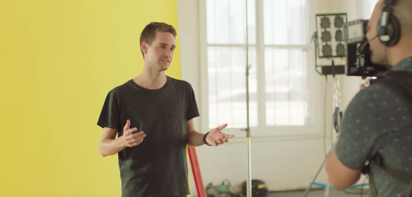 Snap CEO Evan Spiegel stands in front of a yellow background and explains the updated Snapchat design while being filmed by a cameraman