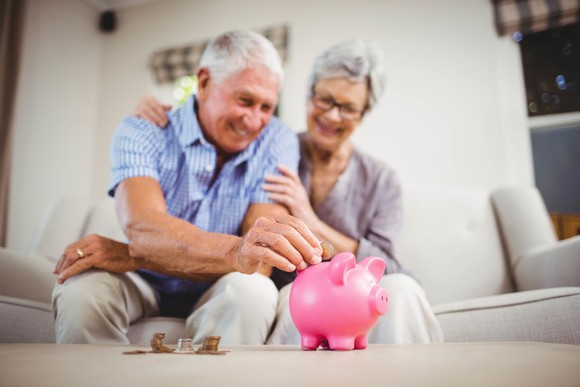 An older man sitting next to an older woman on a couch puts a coin into a piggy bank