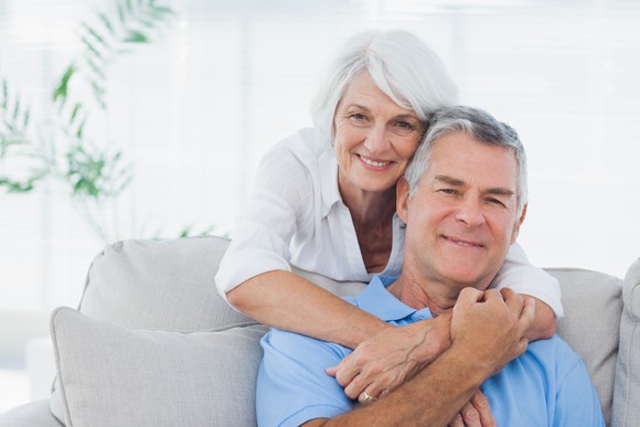 Older couple with man seated on the couch and woman standing with arms around him.