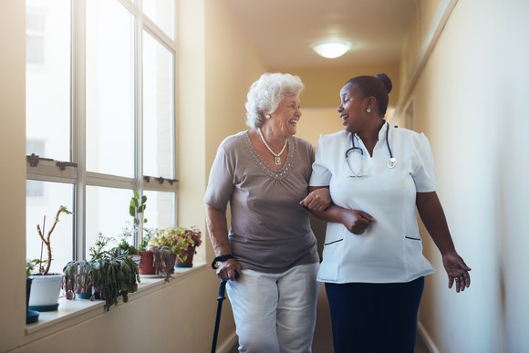 Older-looking person with cane walking with a medical assistant in a corridor with plants and windows.
