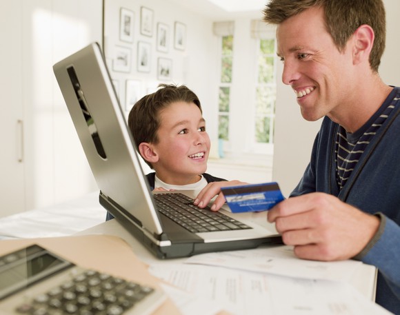 A smiling man holds a credit card and looks at his computer while a boy looks on smiling.