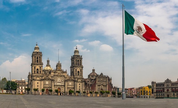 Mexican flag flying over a square with a large palacial building in background.