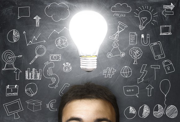 A view of a person's forehead with an illuminated lightbulb above it in front of a background showing a chalkboard covered by diagrams.