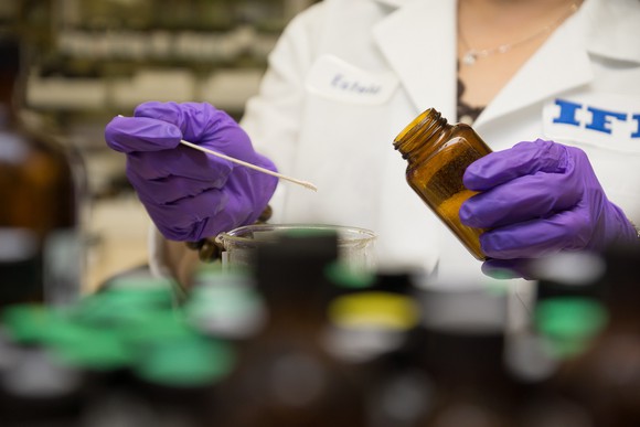 Person in lab coat and purple gloves swabbing contents of a brown bottle into a beaker.