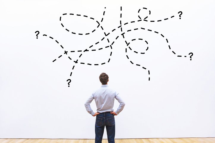 A man staring at a drawing on the wall in front of him of randomly drawn lines ending with question marks.