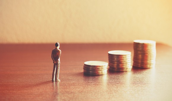 A miniature figurine of a man is standing on a table in front of three stacks of coins.