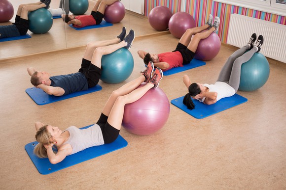 An indoor fitness class showing two young men and two young women each laying on a floor mat with legs raised on a ball.