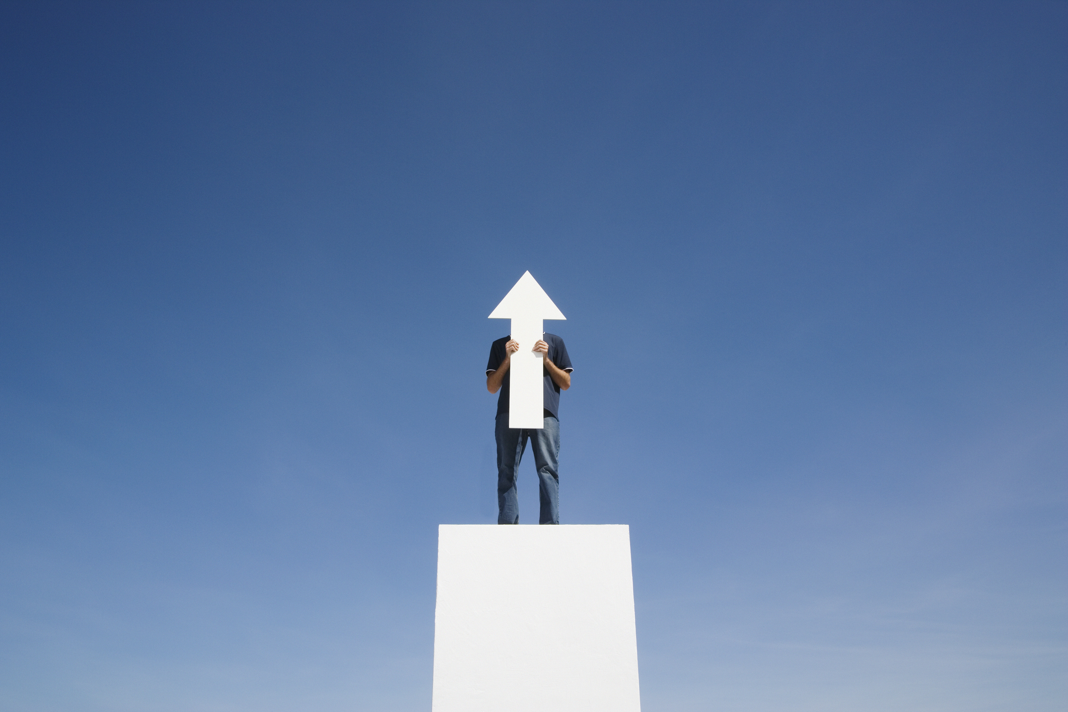 A man standing on a white column against a blue sky and he's holding a large white arrow cutout pointing up.