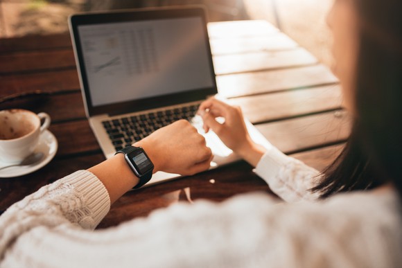 Woman looking at her watch, with an open laptop in front of her next to a cup of coffee