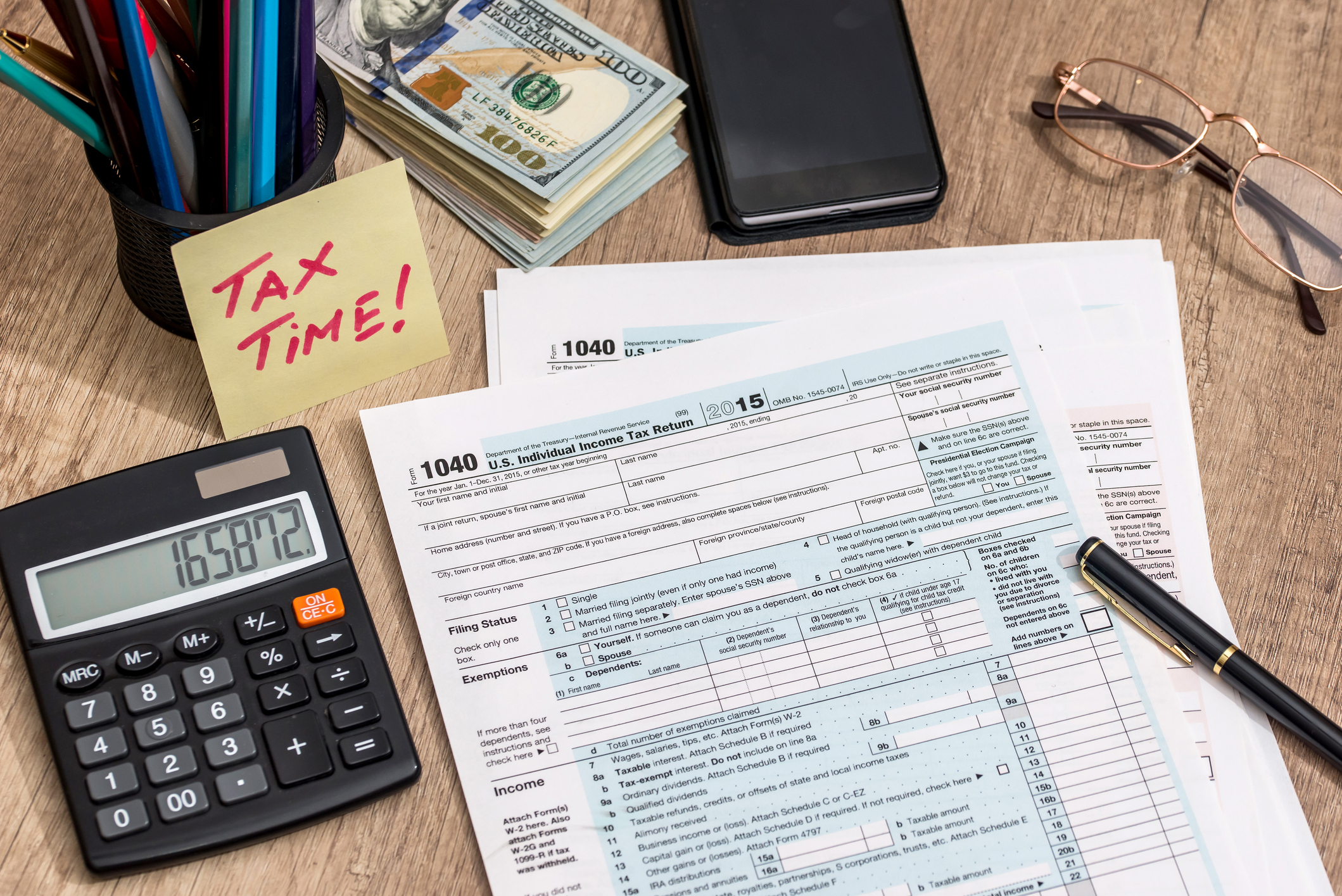 A desktop with a calculator, some pencils, a stack of $100 bills, a pair of glasses, a checkbook, and a note that says tax time next to some tax forms.