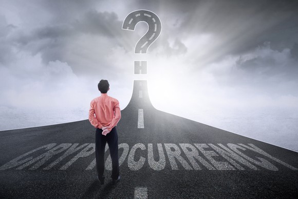 A highway marked with the word Cryptocurrency rises into the sky, ending in a huge question mark. On the blacktop, a man in a red shirt quietly considers the riddles ahead.