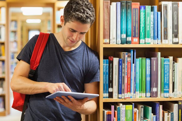 Young male wearing a backpack using a tablet while standing against a library bookshelf