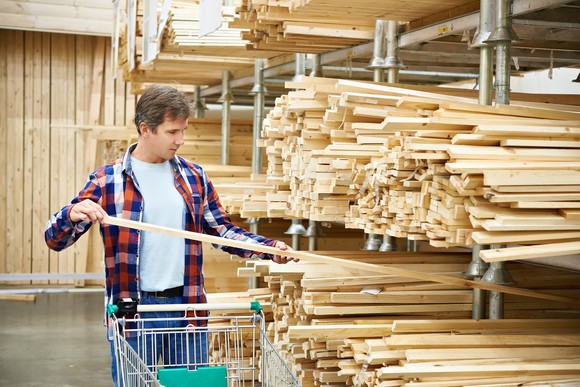 Man with a shopping cart lifting a piece of lumber off of a pile of pieces on a shelf