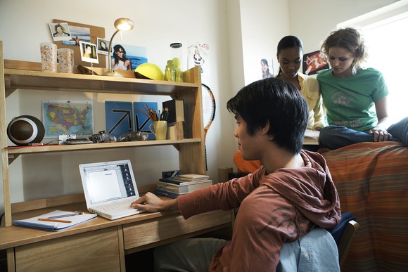 Young male at a laptop with two females sitting on a nearby bed